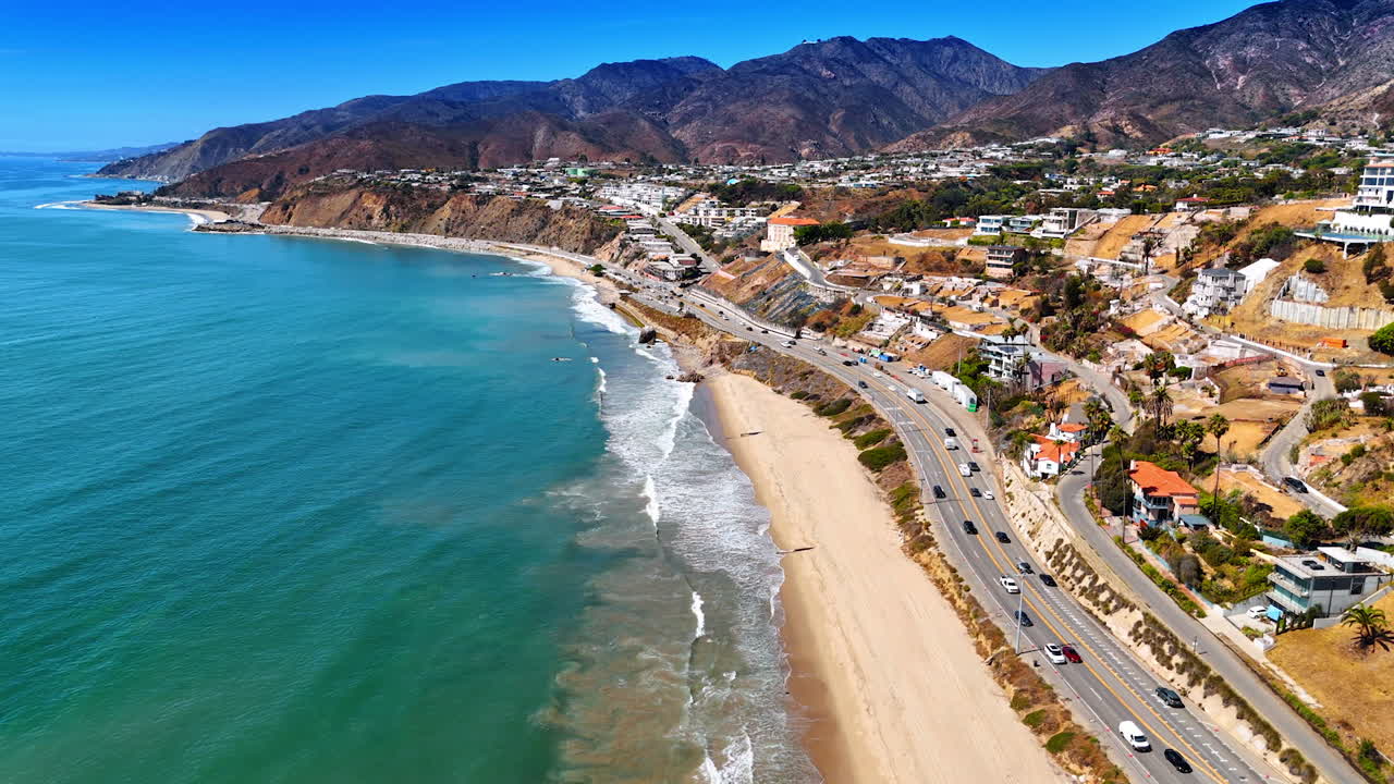 Numerous cars ride by the highway along the sandy beach of the Pacific Ocean. Drone flight along the rocky coast of Malibu, Los Angeles County, California, USA