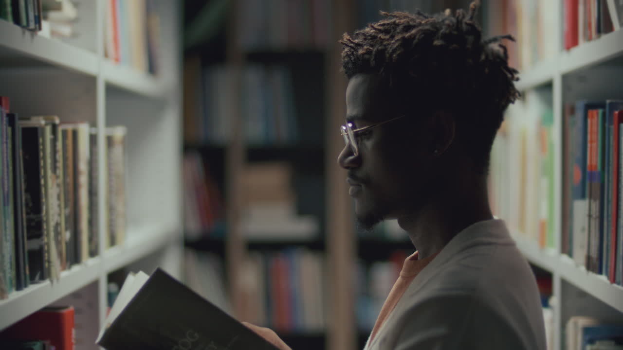 joven afroamericano visitando la biblioteca, leyendo un libro de la estantería