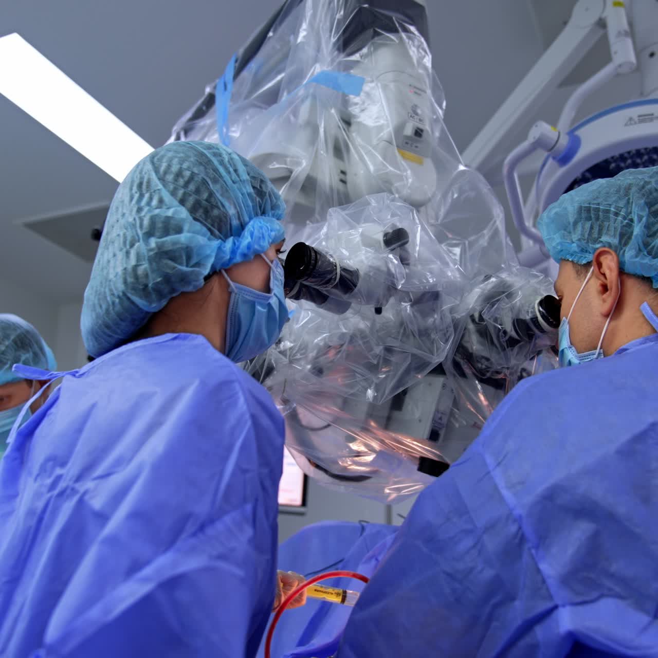 Surgeon and assisting nurse looking into microscope conducting surgery. Male assistant arranging instruments at backdrop