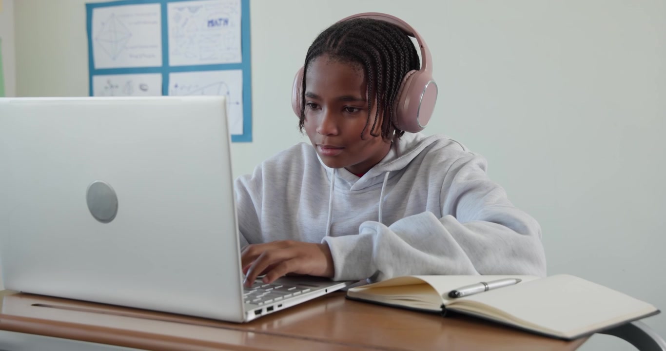 African-American teenage male hooded grabbing pink headset for study at desk with laptop, notebook