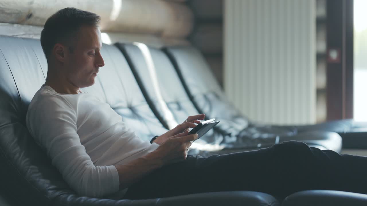 Thoughtful young man is sitting on sofa in room, looking for information on tablet. Side view