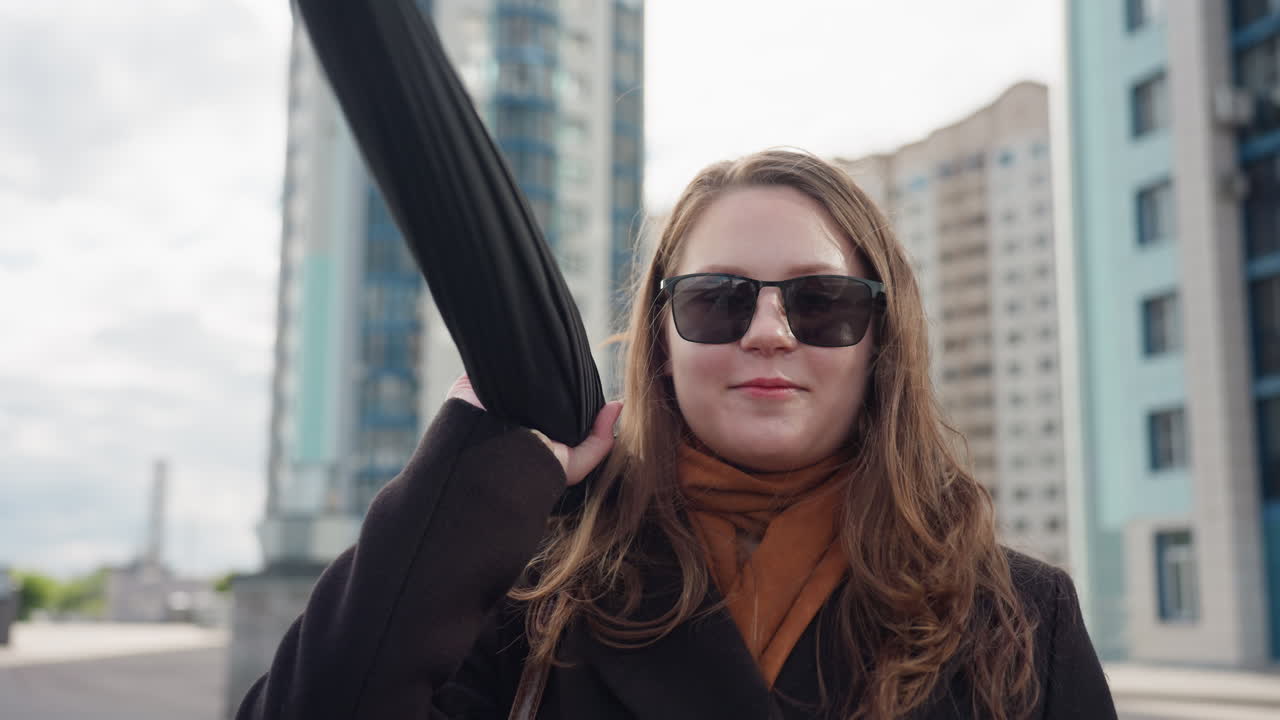 Female student holding folded umbrella tries to throw during city walk, wearing dark long suit, hair moving in wind before modern residential towers, confident stance captured in daylight city scene