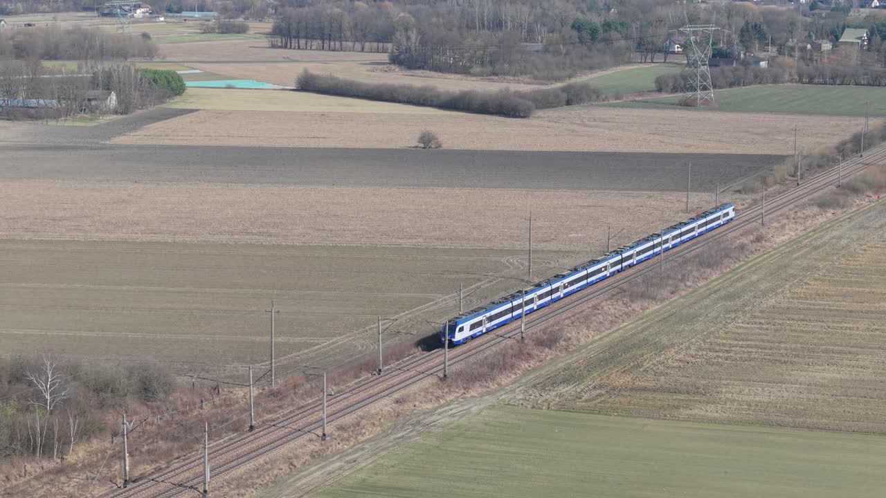 Letecké záběry z dronu moderního osobního vlaku projíždějícího rovinatou venkovskou krajinou s zemědělskými poli a holými zimními stromy. Elektrická železnice s trolejovým vedením protíná úhlopříčně krajinu.