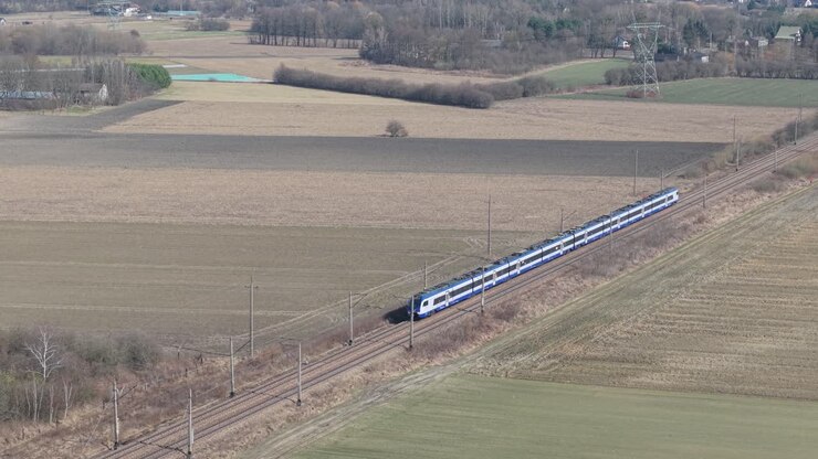 Luchtdronebeelden van een moderne passagierstrein die door een vlak landelijk landschap rijdt met landbouwvelden en kale winterbomen. Elektrische spoorlijn met bovenleidingen snijdt diagonaal.