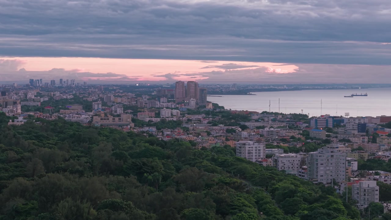 Hochwinkel-Luftaufnahme einer statischen Drohne über dem Stadtbild von Santo Domingo, die das grüne Blätterdach des Mirador Sur Parks und einen dramatischen rosa Dämmerungshorizont über dem Karibischen Meer zeigt.