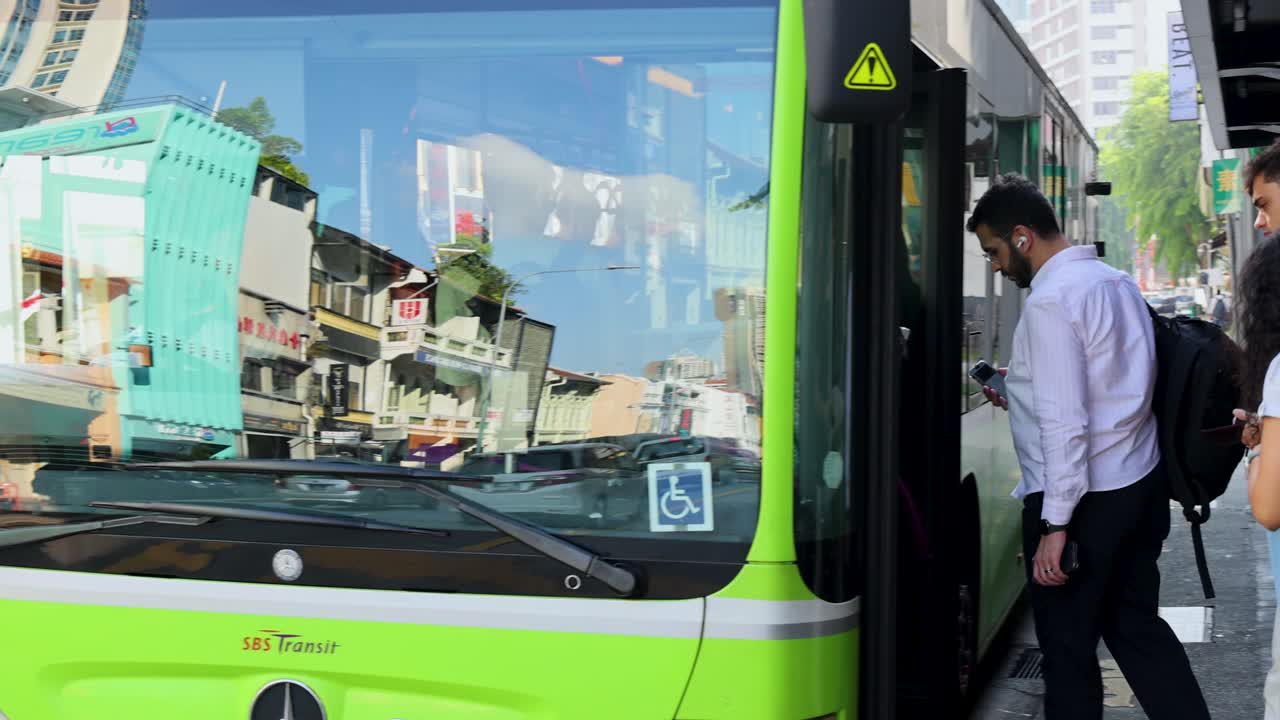 A man in business attire boards a green city bus on a sunny Singapore street, with colorful shophouses reflected in the bus windows. Camera remains steady