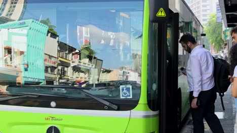 a man in business attire boards a green city bus on a sunny singapore street, with colorful shophouses reflected in the bus windows. camera remains steady