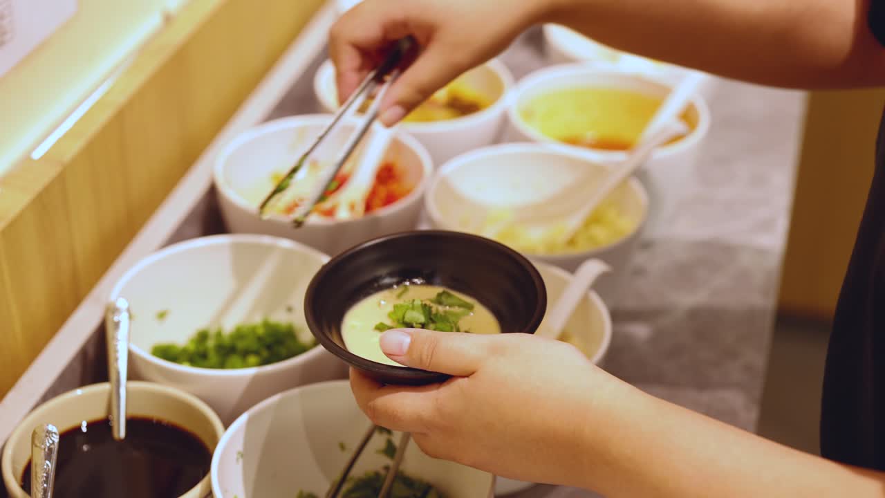 Individual uses chopsticks to add condiments to a bowl at a well-lit sauce station
