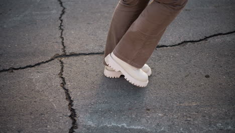 young woman steps in white boots across cracked asphalt pavement under warm sunlight, winter pants swaying in rhythm, tapping echoes through empty urban street lined with melting snow