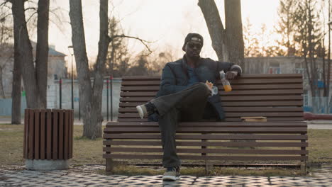 young man sits on park bench eating snack while holding drink, legs crossed, sunglasses and cap on, break at sunset, book on bench, blurred people with stroller in background