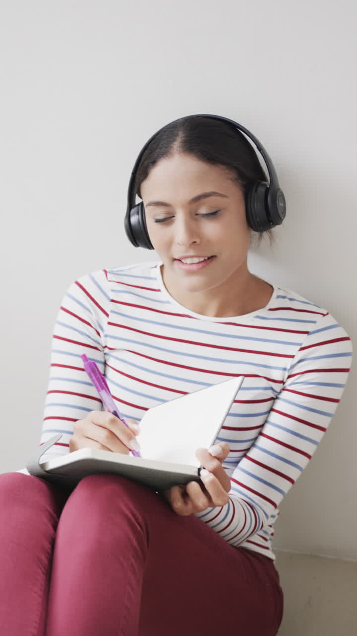 Vertical video of portrait of happy biracial woman using headphones, taking notes, slow motion