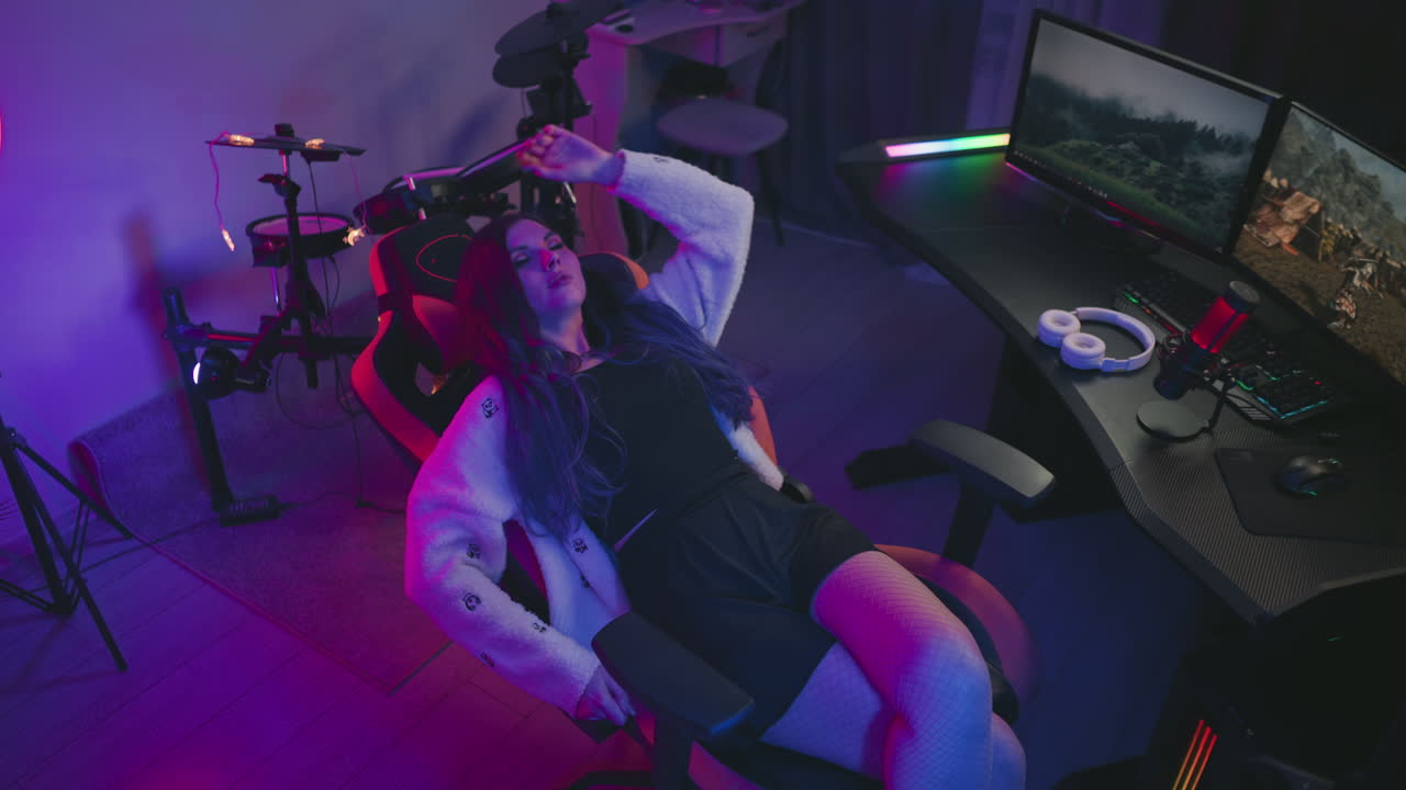 Woman relaxes her gaming chair backward to take break from game, looking down in thoughtful mood, surrounded by dual monitors, drum set, and vibrant LED lighting in cozy modern setup