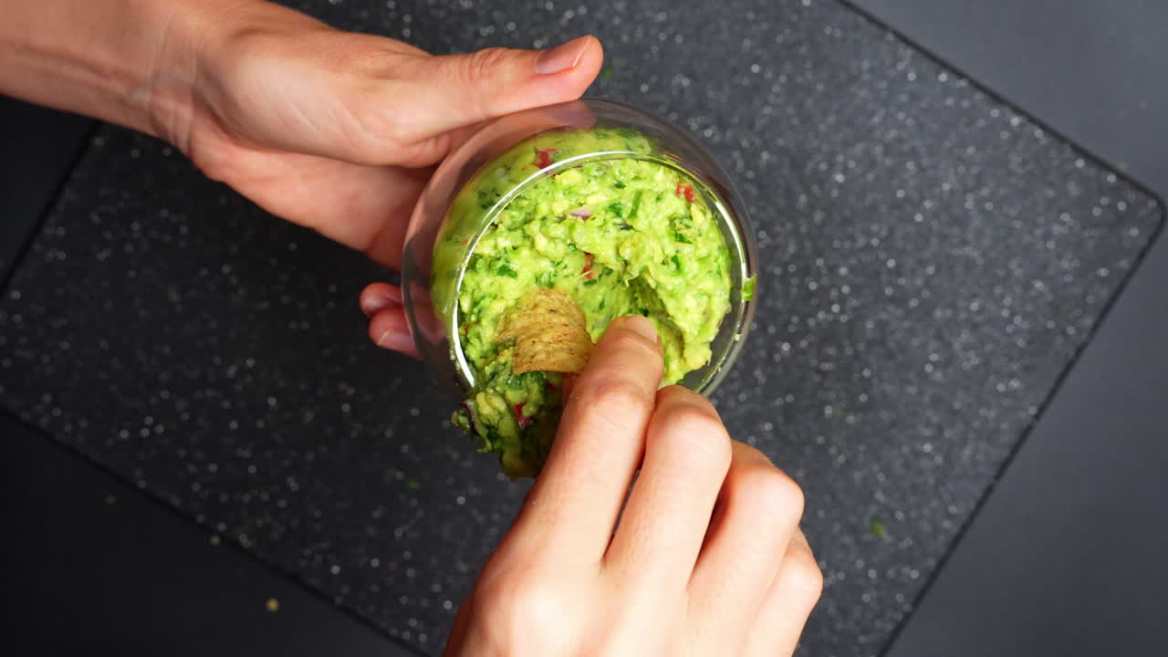 Ruce namáčejí tortilla chips do misky s čerstvým guacamole u kuchyňského stolu. Dvě ruce drží tortilla chips a namáčejí jej do misky se zeleným guacamole v kuchyni.