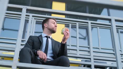 Happy Young Banker Sitting on the Background of a Modern Building and Eating a Tasty Hot Dog
