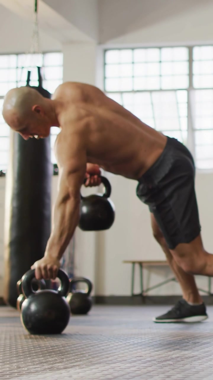 Vertikal video: Man greppar kettlebells på ett gym, utför alternerande roddövningar för att bygga styrka.