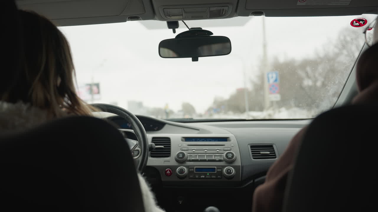 Behind view of people inside moving car with driver holding steering wheel and passenger seated beside while road signs, trees, buildings, and cloudy sky are visible through windshield during daytime
