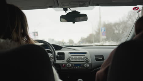 behind view of people inside moving car with driver holding steering wheel and passenger seated beside while road signs, trees, buildings, and cloudy sky are visible through windshield during daytime