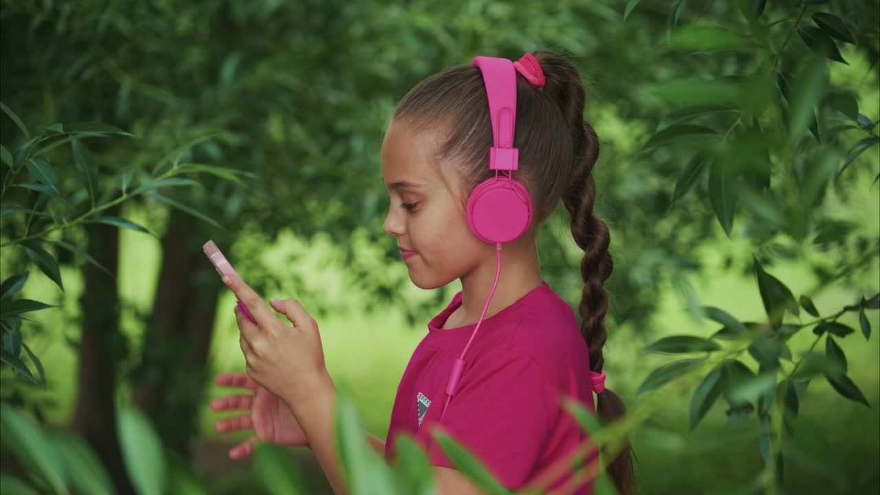 A young girl enjoying her music with pink headphones, immersed in nature, using a smartphone while surrounded by greenery, showcasing a moment of joy and connection