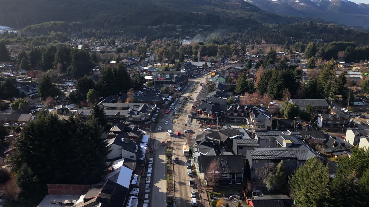 Aerial fly over the mountainous Villa La Angostura town main road lined by buildings and trees, Neuquén, Argentina