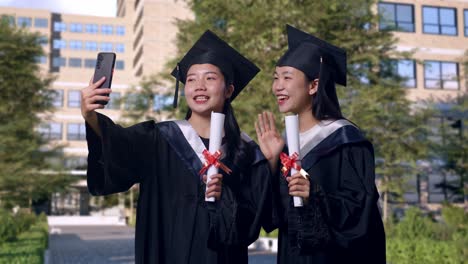 asian woman students graduate in caps and gowns with diplomas having a video call on smartphone while standing in front of a magnificent university building