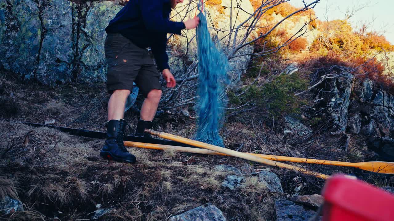 A Man Prepares a Blue Fishing Net Beside a Rocky Outcrop, With Wooden Oars Lying Nearby Near Lake Reinsjøen in Åfjord, Trøndelag, Norway - Static Shot