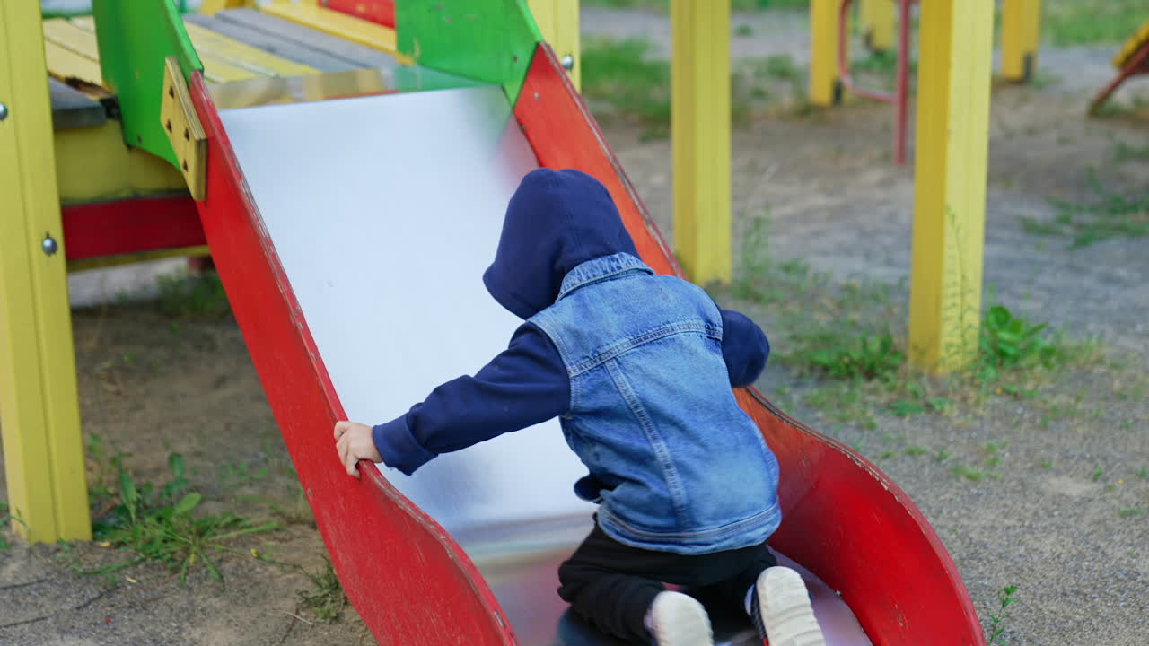 Baby boy wearing a jacket with a hood tries to climb up the slide. Kid fails and walks away.