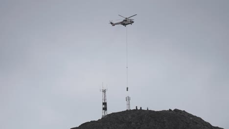 un poderoso helicóptero ayuda a instalar equipos en la cima de una montaña, con una torre de comunicaciones cerca