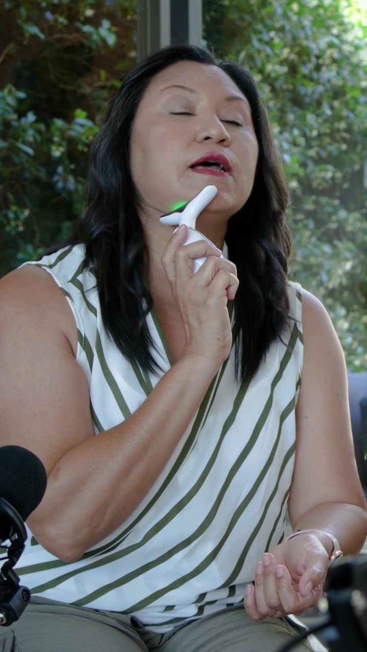 Video verticale: Donna che mostra l'uso di un dispositivo facciale a casa, con un microfono e un top a righe.