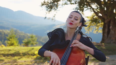 mid-aged young woman with makeup and nice black dress performs cello music. lady closed her eyes enjoying music. nature backdrop in blur.