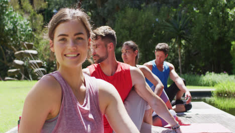 mujer caucásica sonriente practicando yoga con un grupo diverso sentada en un parque soleado