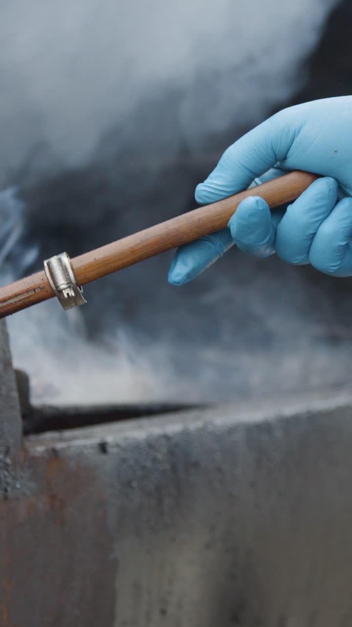 A person wearing blue gloves uses a wooden spatula to flip burgers on a smoky, outdoor metal grill. Natural lighting, close-up, steady camera
