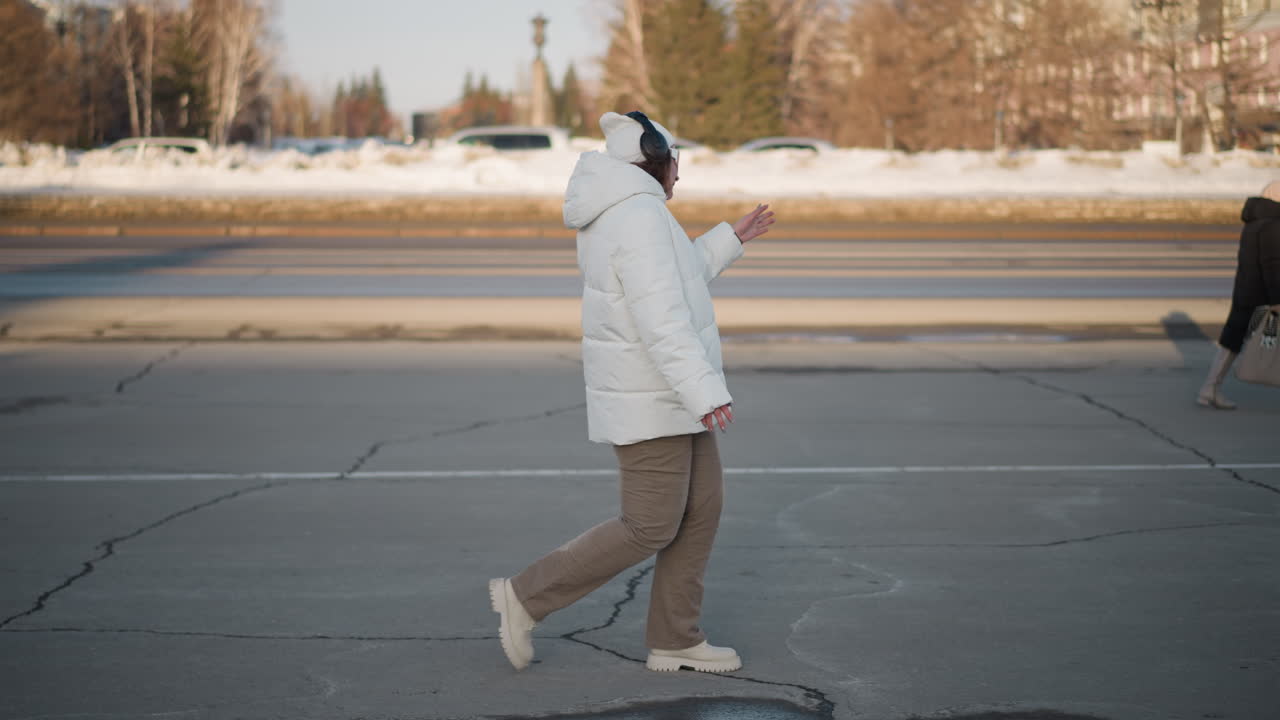Side view of figure in white winter coat and sunglasses dancing joyfully on cracked pavement, snowy background and passing vehicles behind, expressing happiness and freedom on cold city street
