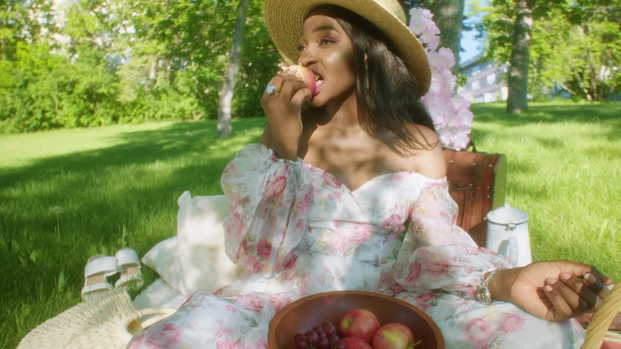 mujer negra sonriente en el parque en un picnic con vestido floral le da un mordisco a la manzana