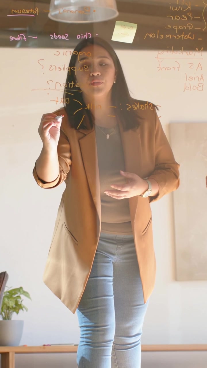 Video verticale: Sollevando un pennarello, una donna con blazer color cammello e jeans scrive per la pianificazione su una lavagna di vetro