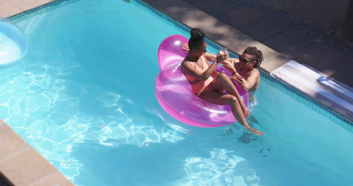 Due donne che arrivano galleggiando su un materassino rosa nella piscina di casa, passandosi una bevanda, socializzando, con spazio per il testo