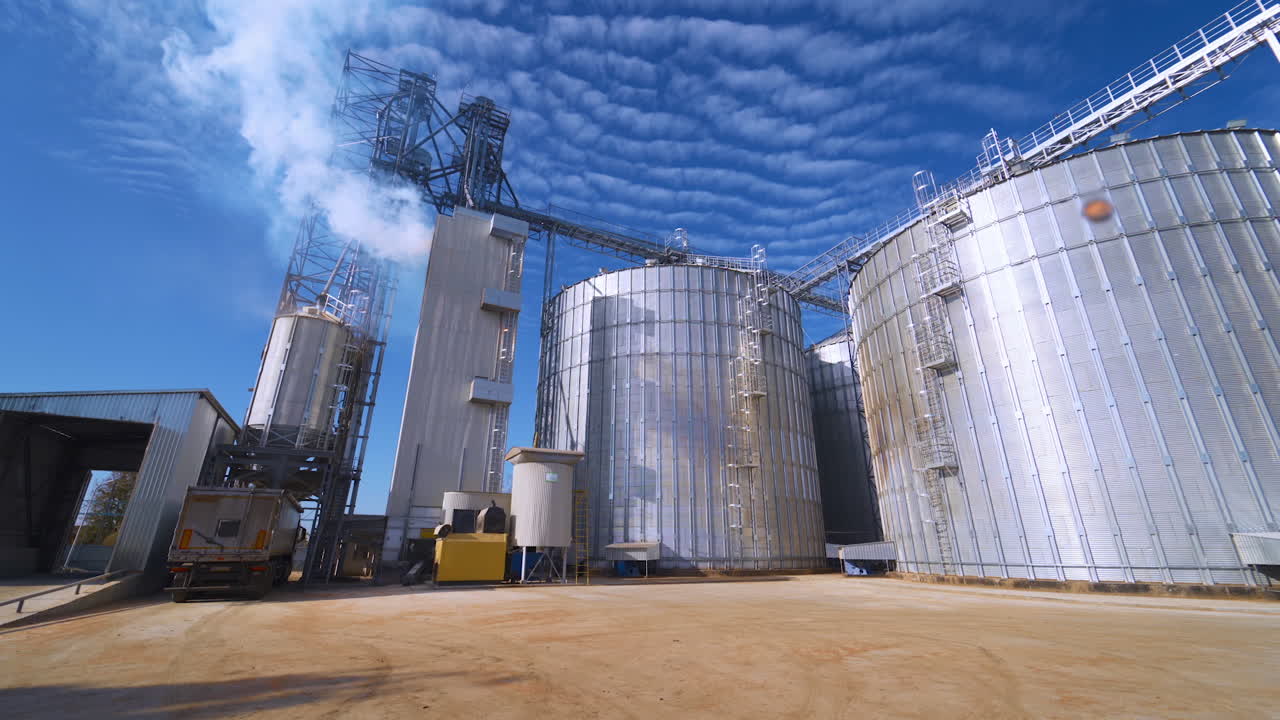 Modern factory under blue sky. Exterior of large grain elevators at sunlight. White vapor goes into the air from industrial plant.