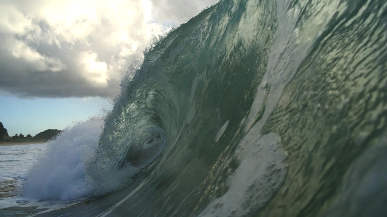 Curling green ocean wave rises beside whitewater under cloudy sky in fluid slow motion
