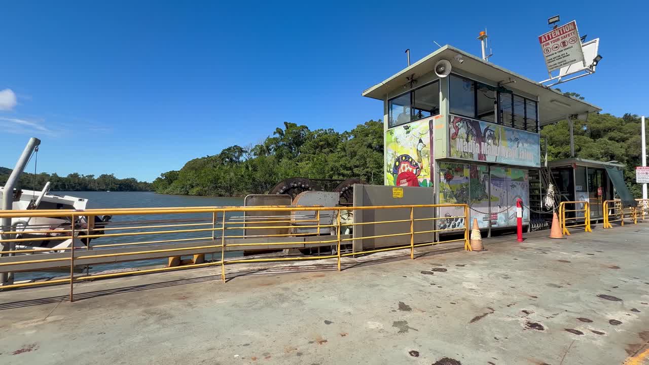 A cable ferry with vehicles crosses the Daintree River under clear daylight, passing a control tower with painted murals. Camera pans steadily across the scene