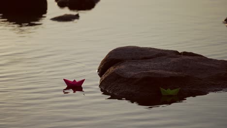 paper boats in water over beautiful sunset. two colorful paper ships floating on river surface near the big stone. origami ship sailing.