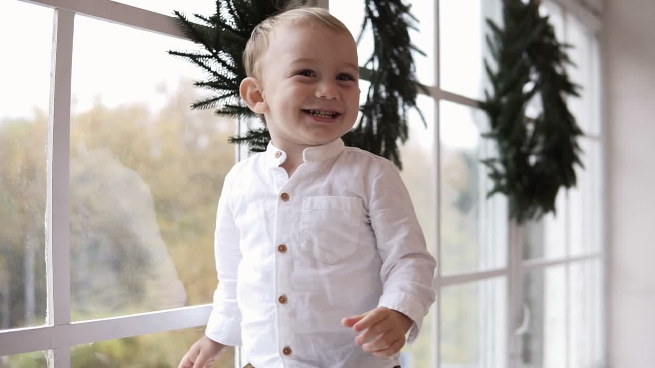 Cute baby boy in white shirt standing on the window sill and smiling. The window is decorated with Christmas wreath