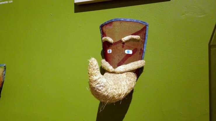 Primo piano di maschera decorativa artigianale con barba intrecciata, montata su parete museale, che mostra arte popolare tradizionale, artigianato testurizzato e design culturale