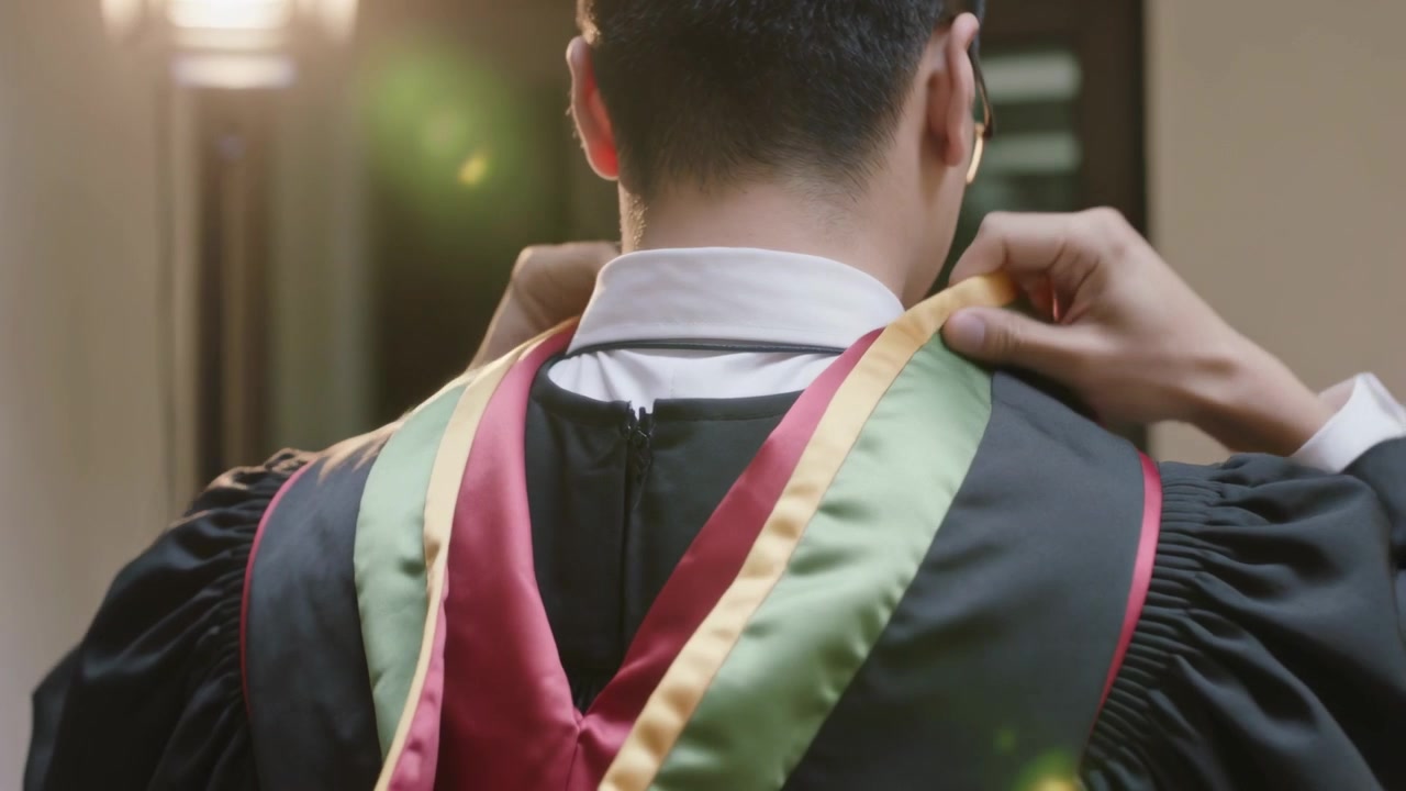 Laureato che si sistema il cappuccio rosso e verde sopra la toga nera in un corridoio per la laurea, con microfono.