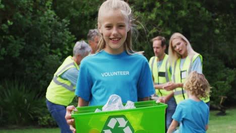 Smiling-caucasian-girl-holding-recycling-box-picking-up-litter-with-volunteers-in-field