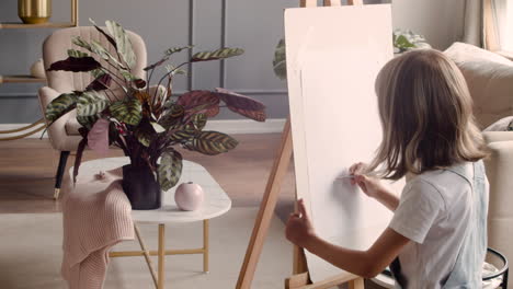 rear view of little blonde girl painting on the lectern next to the window at home