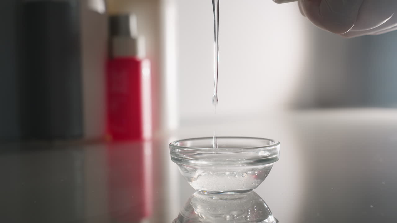 Bodywork therapist wearing gloves pours clear massage oil drop by drop into small white bowl on reflective trolley surface in softly lit spa room, highlighting calm precision and professional care