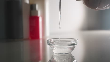 bodywork therapist wearing gloves pours clear massage oil drop by drop into small white bowl on reflective trolley surface in softly lit spa room, highlighting calm precision and professional care