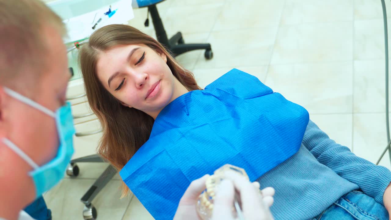 Beautiful young girl smiles and listens to the dentist. The doctor shows the layout of the jaw