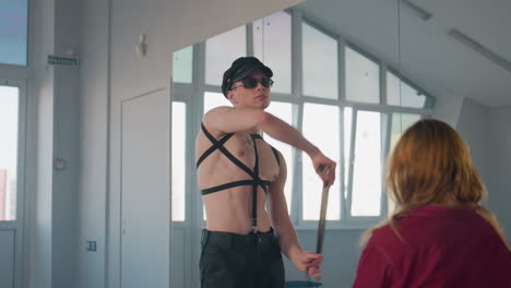 rear view of woman with golden hair seated facing man removing belt looping it around neck then casting aside during seductive dance in mirrored room spotlighting harness sunglasses baton cloth props