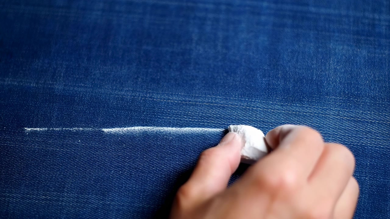 Close view shows hand pressing white chalk against blue denim, creating bright marking line across textured weave while tailoring work, repair, and garment planning feel precise, with hand, chalk