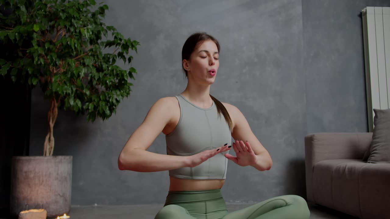 Zoom-in Confident brunette girl in a gray top and green sweatpants does yoga sits in the lotus position and practices breathing practices in a modern apartment at home near a houseplant